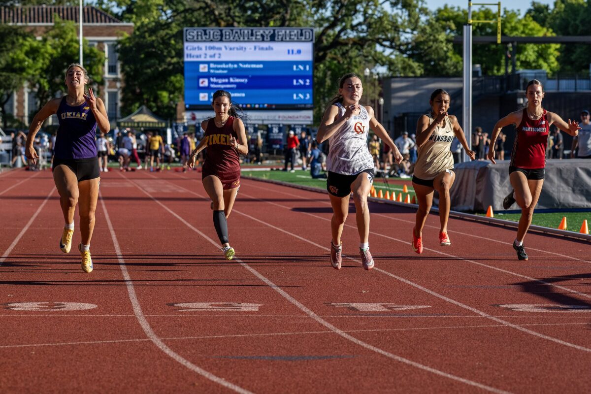 Five runners approaching finish beneath scoreboard